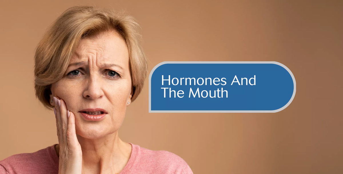A woman with short blonde hair rests her hand on her cheek, looking distressed, with a blue text label reading 'Hormones And The Mouth' on a warm brown background.