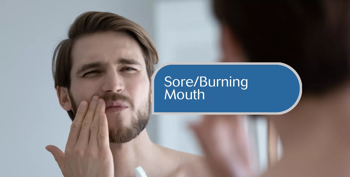 A man in a bathroom looking into a mirror, touching his cheek as if feeling a sore or burning mouth, with a blue text overlay that reads 'Sore/Burning Mouth'.